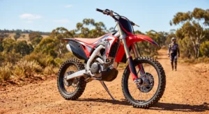 A high-performance red and white dirt bike resembling a Honda CRF450R is parked on a red dirt trail in a dry Australian outback landscape, with a blurred rider standing in the distance under a blue sky.